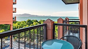 Un balcón con una mesa de vidrio y sillas de mimbre, con vistas a una piscina y un paisaje con mar y montañas.