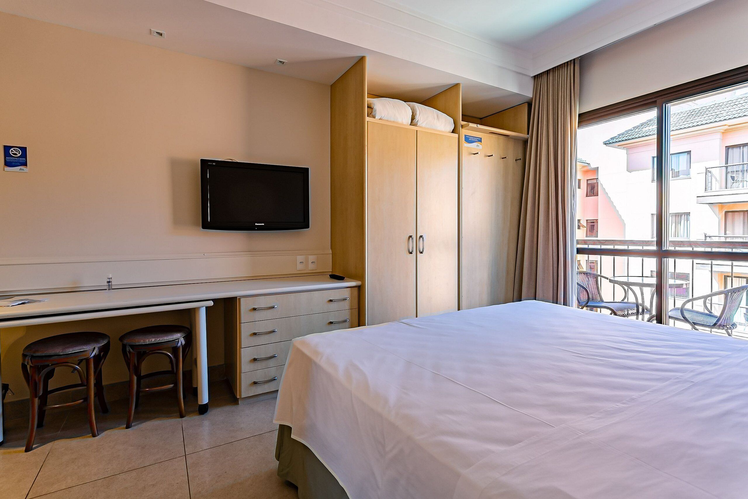 A hotel room with a bed, a desk with two stools, a television, and a wardrobe.