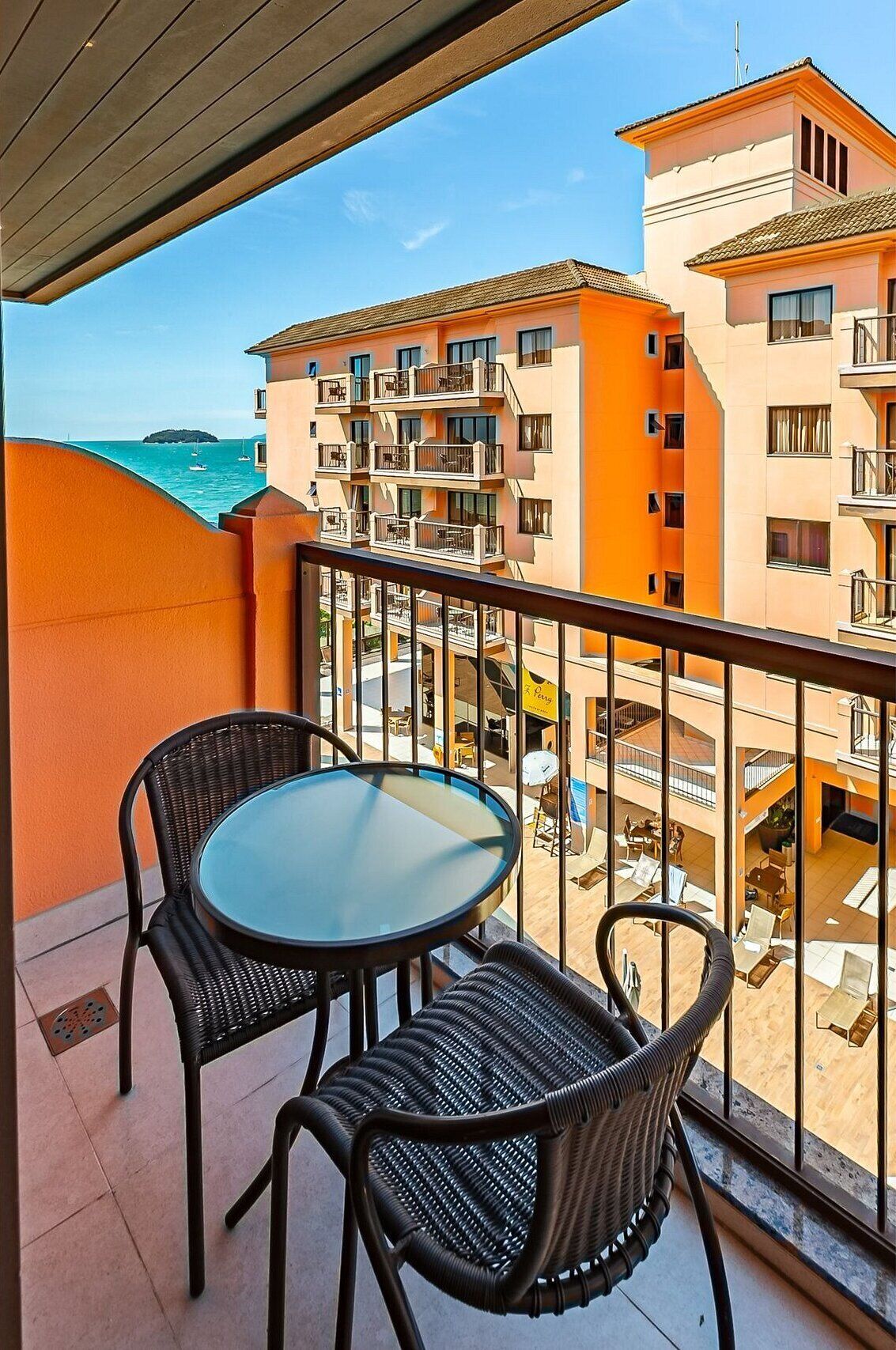 A balcony with two wicker chairs and a glass table, overlooking a building with multiple balconies and a view of the sea.