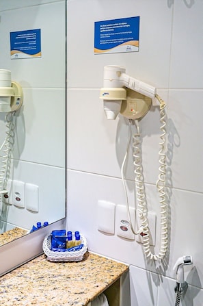 Een badkamer met een haardroger aan de muur, een spiegel, een wastafel met toiletartikelen en een handdoekhouder.