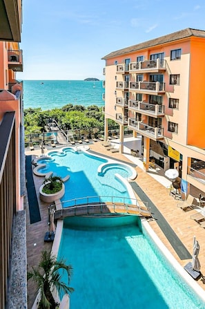 Een zwembad van een resort met uitzicht op de zee, omringd door een meerlaags gebouw met balkons.