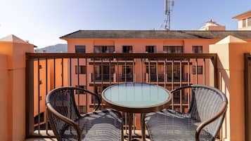 A balcony with two wicker chairs and a glass table, overlooking a building with multiple windows and balconies.