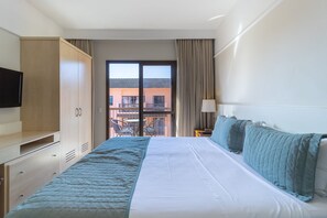 Ein Hotelzimmer mit Bett, Fernseher, Kleiderschrank, einem Balkon mit Stühlen und Blick auf Gebäude.