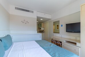 Quarto de hotel com cama, escrivaninha, televisão, cozinha compacta e banheiro visível através de uma porta aberta.