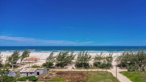 A coastal view with a sandy beach, a clear blue sky, and a few buildings in the foreground. - Loft 50 m From Camacho Beach Plo0002 (Jaguaruna)