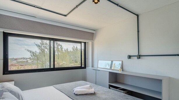 A modern bedroom with a large window, a bedside table with a towel, and a built-in shelf. - Loft 50 m From Camacho Beach Plo0002 (Jaguaruna)