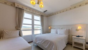 Een hotelkamer met twee bedden, een groot raam met gordijnen, een nachtkastje met een lamp en uitzicht op zee.