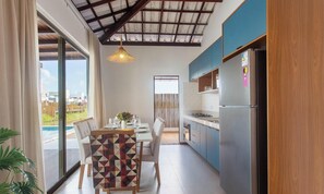 A modern kitchen with a large island, a dining area with a geometric-patterned chair, and a view of a swimming pool through large windows. - 2-suite House by the Lagoon in Marau Cdl0008 (Maraú)