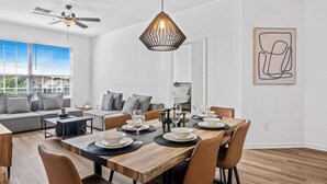 A modern living room with a dining area, a ceiling fan, and a dining table set for four. - Beautiful Condo at Windsor Palms Resort (Kissimmee)