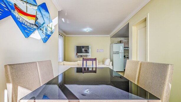 A dining area with a glass-top table and cushioned chairs, a television, and a refrigerator in the background. - House With Pool in Cond 150m Beach Mariscal Ccm004 (Bombinhas)