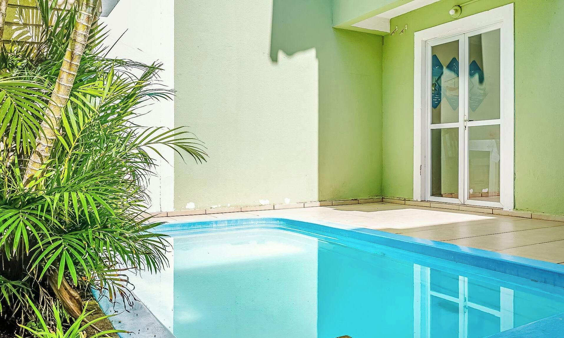 Ein Schwimmbecken mit klarem, blauem Wasser, umgeben von einer gefliesten Terrasse, neben einem Gebäude mit grüner Fassade und einer Glastür.