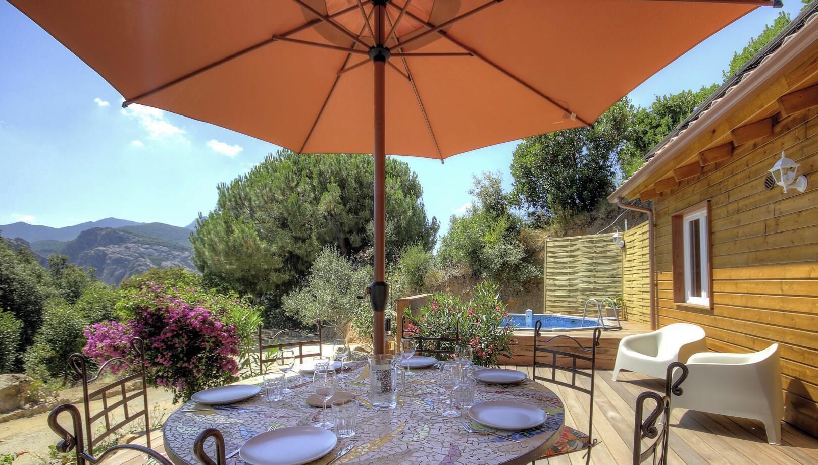 A round table with a mosaic tablecloth, surrounded by chairs, under a large umbrella, with a pool and wooden cabin in the background.