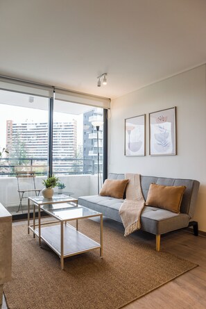 A modern living room with a grey sofa, a glass coffee table, and a view of a cityscape through large windows. - Beautiful Apt in Las Condes with Pool (Las Condes)