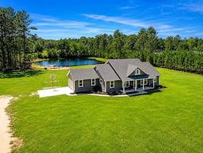 A single-story house with a covered porch, a lawn, and a pond in the background.