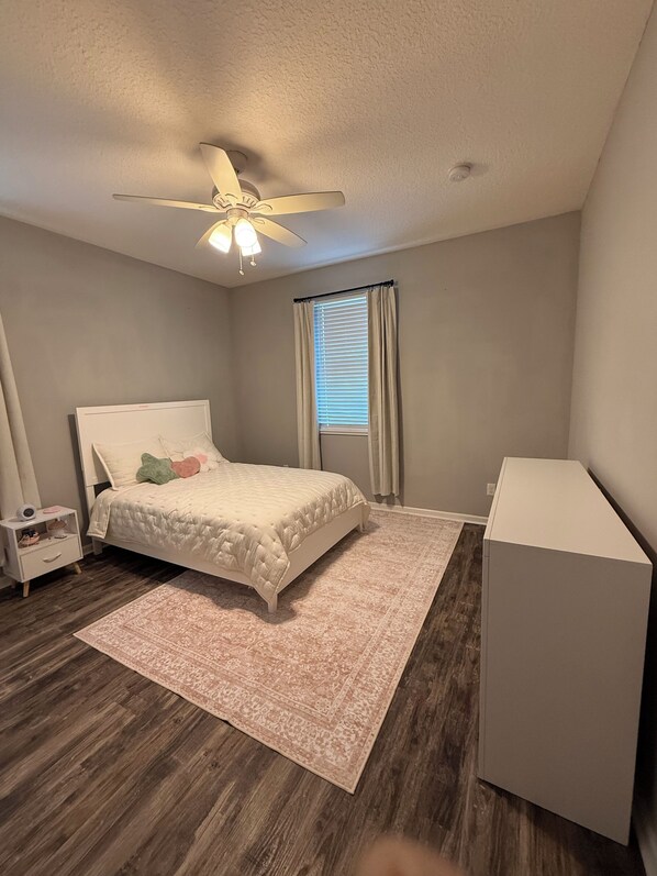 A bedroom with a bed, a nightstand, a desk, a ceiling fan, and a window with curtains.