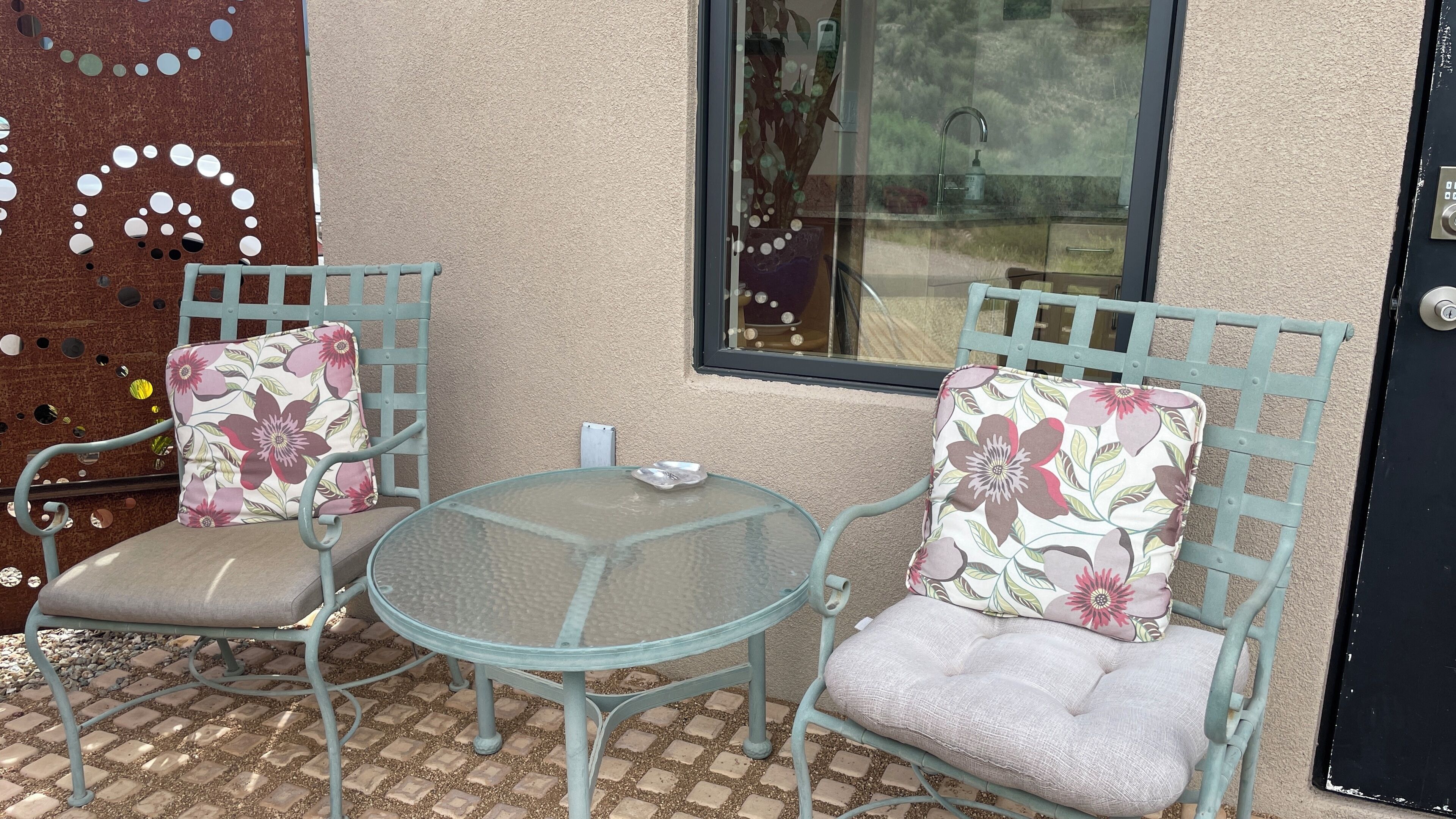 Un petit espace extérieur aménagé avec deux chaises et une table, une fenêtre donnant sur un paysage agréable et une porte dotée d’un système de verrouillage à clavier.