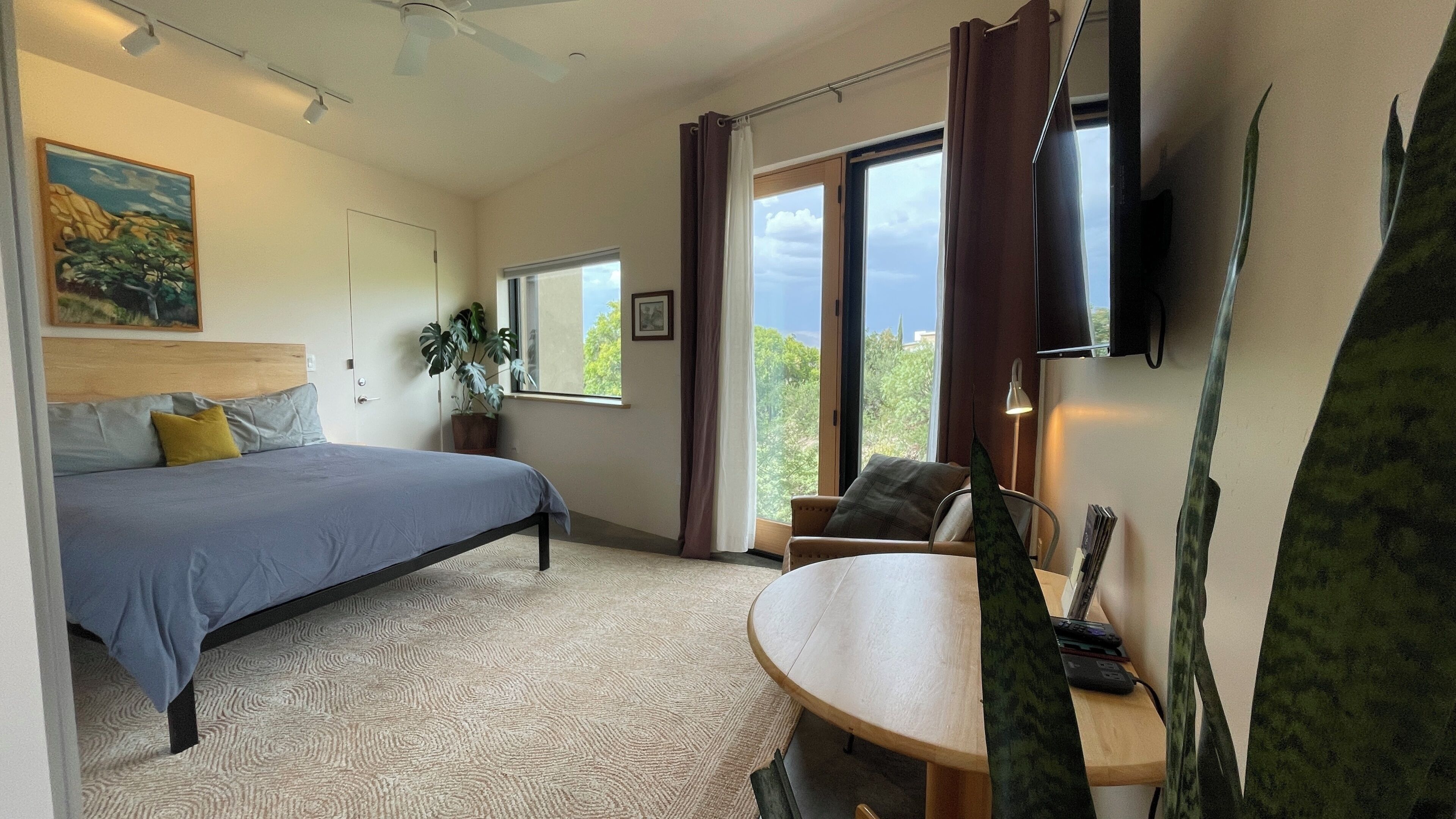 A bedroom with a bed, a desk, a chair, a plant, and a window with a view.