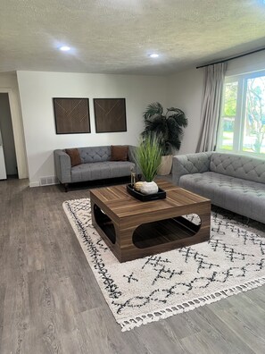 Ein modernes Wohnzimmer mit einem Holztisch, grauen Sofas und einem großen Fenster mit Vorhängen.