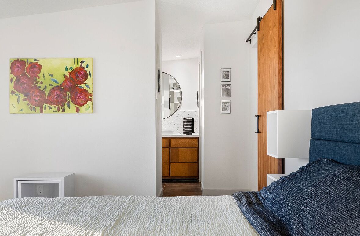 Une chambre à coucher avec un lit, une table de chevet, une lampe, un tableau et une vue sur la cuisine.