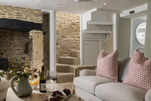 Ein gemütliches Wohnzimmer mit Steinwand, Kamin, einer Couch mit gemusterten Kissen und einem Tisch mit einer Weinflasche und Trauben.