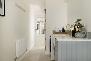 A hallway leading to a bathroom with a white radiator, a door with a stone wall, and a vanity with a vase of flowers. - 4 Wraggs Row -  a cottage that sleeps 6 guests  in 3 bedrooms (Stow-on-the-Wold)