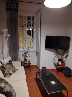 A living room with a sofa, a coffee table, a TV mounted on the wall, a window with a decorative grille, and a potted plant. - Apartment near Feira da Ladra & Alfama (Lisboa)