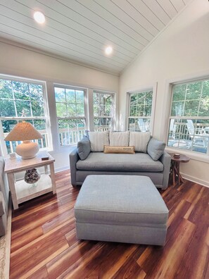 A room with a grey sofa, a matching ottoman, a small table with a lamp, and large windows with a view of trees. - The Tree House (Atlantic Beach)