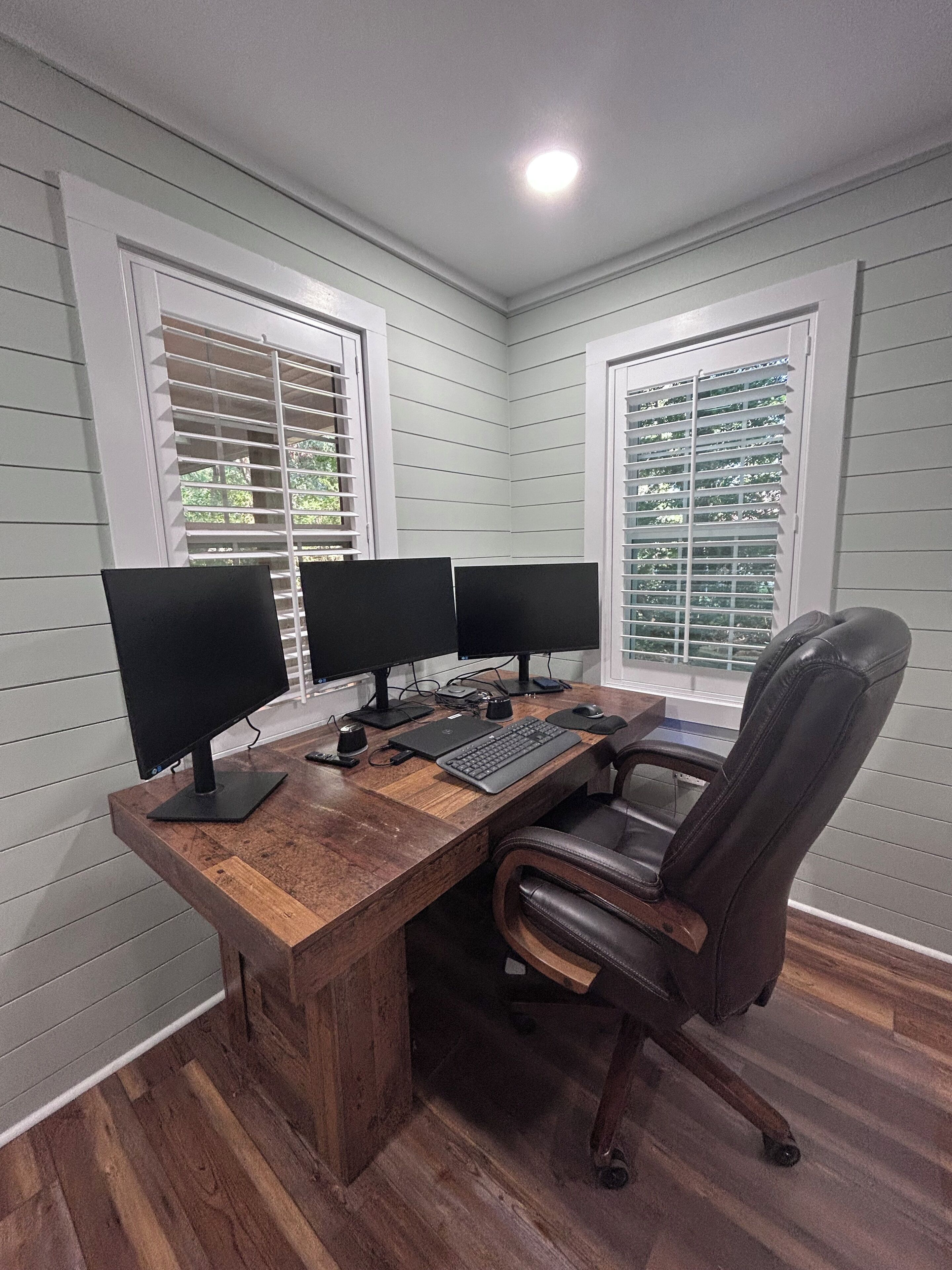 A corner office with a wooden desk, two monitors, a keyboard, and a leather chair.