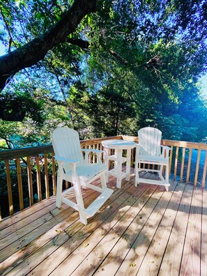A wooden deck with white chairs and a small table, surrounded by lush greenery and trees. - The Tree House (Atlantic Beach)