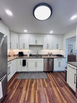 A modern kitchen with white cabinets, a stainless steel refrigerator, a dishwasher, and a patterned rug. - The Tree House (Atlantic Beach)