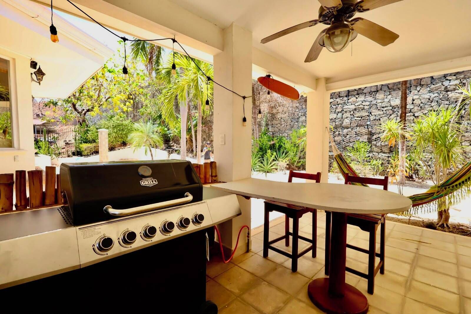 An outdoor patio with a grill, dining table, chairs, a hammock, and a view of a garden and stone wall.
