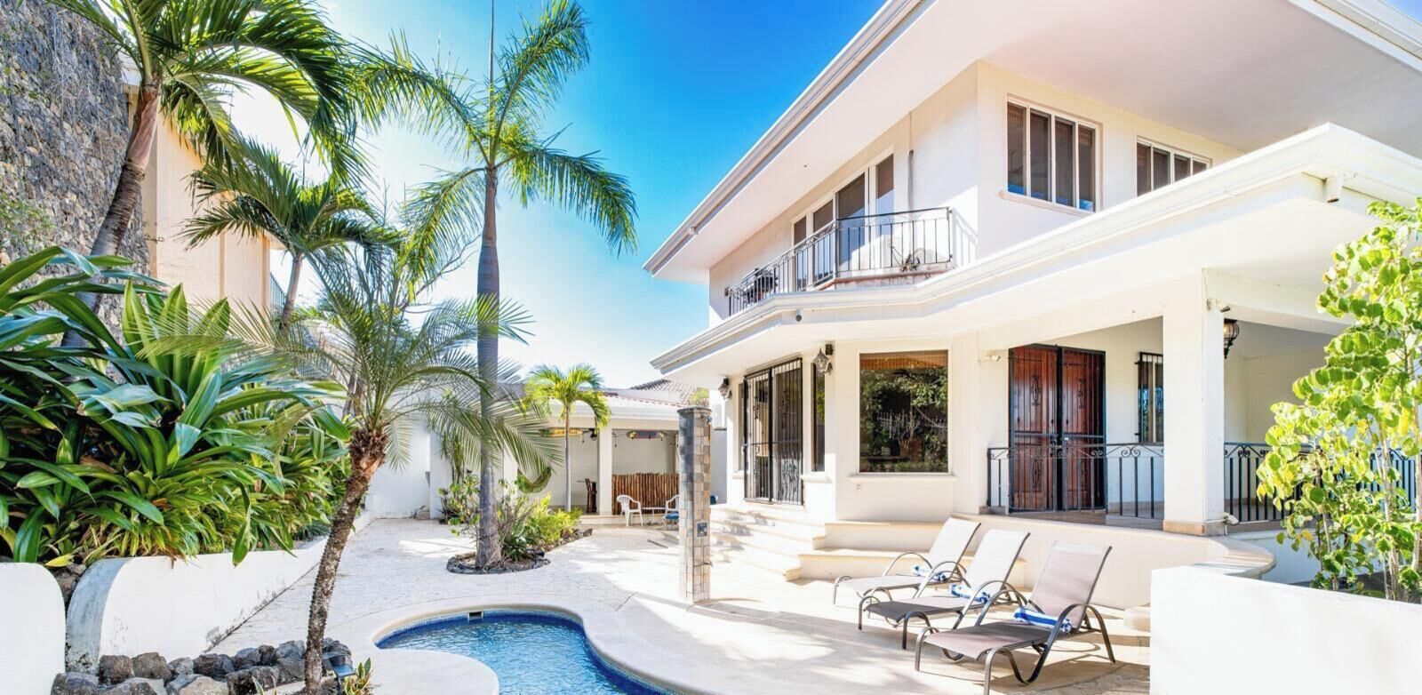 A two-story house with a swimming pool, outdoor seating, and tropical plants.