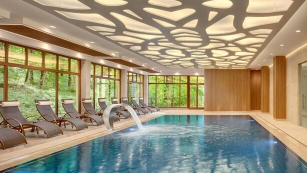 An indoor swimming pool with a waterfall feature, surrounded by lounge chairs and large windows offering a view of greenery. - Svoboda Ensana Health Spa Hotel (Marianske Lazne)