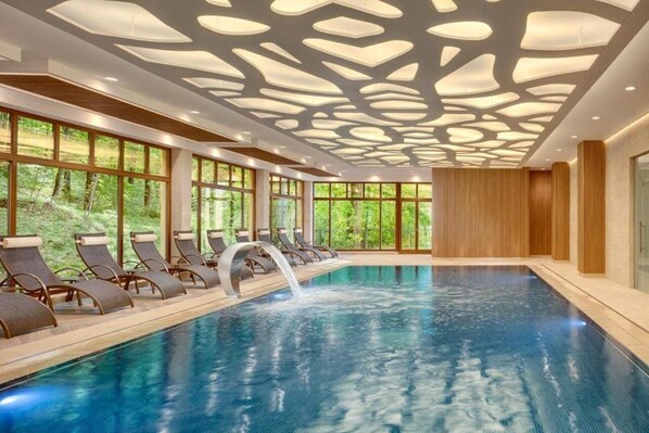An indoor swimming pool with a waterfall feature, surrounded by lounge chairs and large windows offering a view of greenery. - Svoboda Ensana Health Spa Hotel (Marianske Lazne)