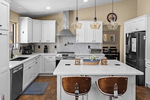 A modern kitchen with white cabinetry, a central island with bar stools, a stainless steel refrigerator, and a tiled backsplash. - Lakefront Retreat | Free Heated Pool + Game Room (Glendale)