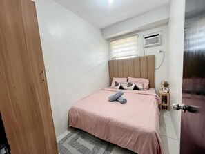 Ein Schlafzimmer mit Bett, Nachttisch, Fenster mit Jalousien, Klimaanlage und Tür.