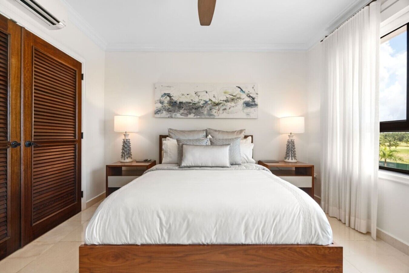 A bedroom with a bed, two bedside tables, a ceiling fan, a painting, and a window with curtains.