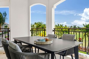 A balcony with a table and chairs, a view of palm trees, and a building in the background. - Luxury Oceanview Condo | Hyatt PR Golf & Beach (Rio Grande)