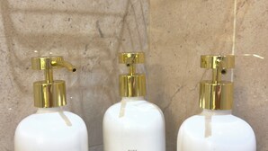 Three white bottles with gold pumps on a gold wire shelf against a marble wall.