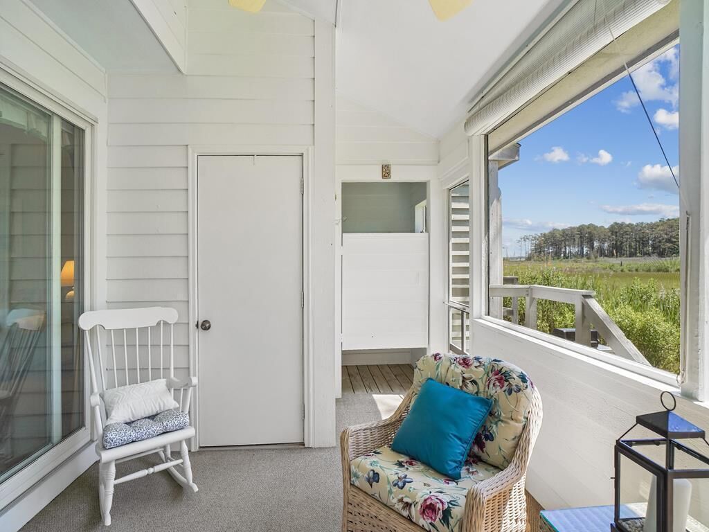 Een overdekte veranda met een witte deur, een rieten stoel met een kussen, en uitzicht op een groen landschap.