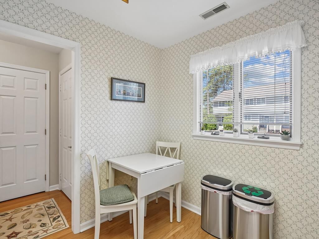 Une pièce avec une table blanche, une chaise, une fenêtre avec des stores, une porte et deux poubelles.