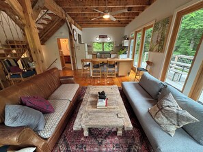 A cozy living room with a wooden ceiling, a dining area with a kitchen in the background, and a large window offering a view of the outdoors.