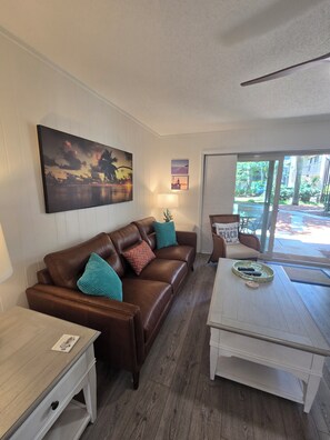 A living room with a brown leather sofa, a white coffee table, and a painting on the wall. - First Floor Flat, Surf Court Villa, 2-King Beds, Steps to Coligny Beach! (Hilton Head Island)