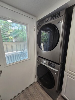 A compact laundry room with a front-loading washer and dryer, a door with a window, and a view of the outside. - First Floor Flat, Surf Court Villa, 2-King Beds, Steps to Coligny Beach! (Hilton Head Island)