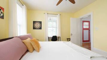 Une chambre d’hôtel avec un lit, un ventilateur de plafond, une fenêtre avec des rideaux, une porte et une petite table avec une chaise.