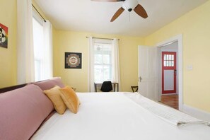 A hotel room with a bed, a ceiling fan, a window with curtains, a door, and a small table with a chair. - Gville and Chill House (Gainesville)