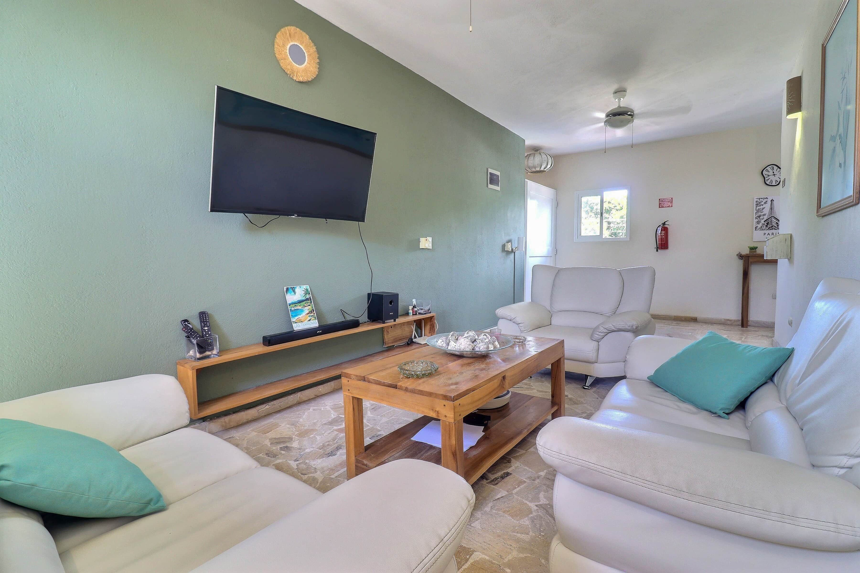A living room with a flat-screen TV mounted on the wall, a wooden coffee table, white sofas with teal cushions, and a stone-tiled floor.