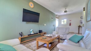 A living room with a flat-screen TV mounted on the wall, a wooden coffee table, white sofas with teal cushions, and a stone-tiled floor.