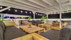 An outdoor seating area with wooden tables and sofas, string lights, and a view of the night sky.