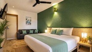A hotel room with a bed, a sofa, a wall-mounted TV, a ceiling fan, and a bedside table with a lamp.
